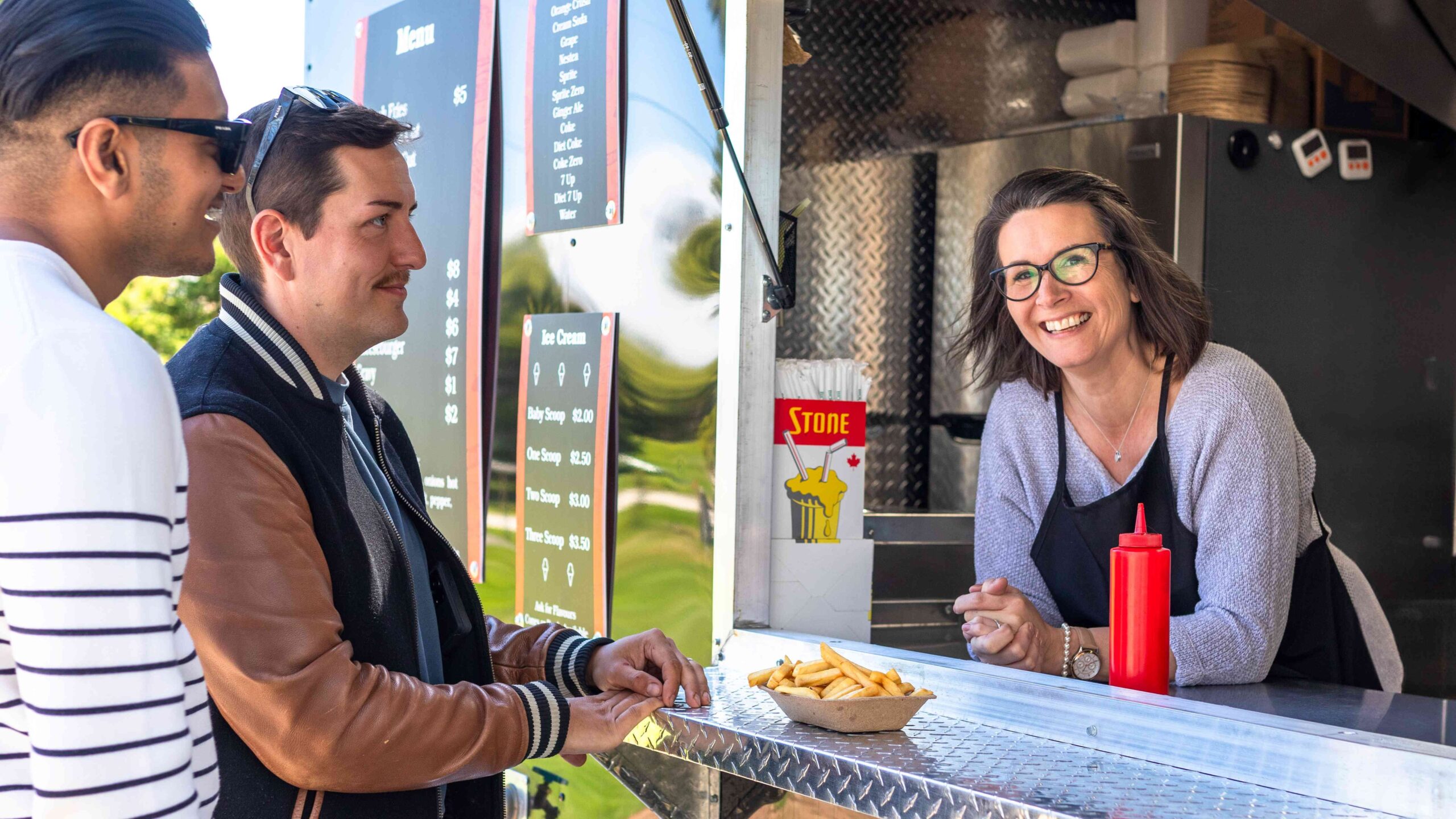 A woman smiles from a food truck service window while serving two customers.