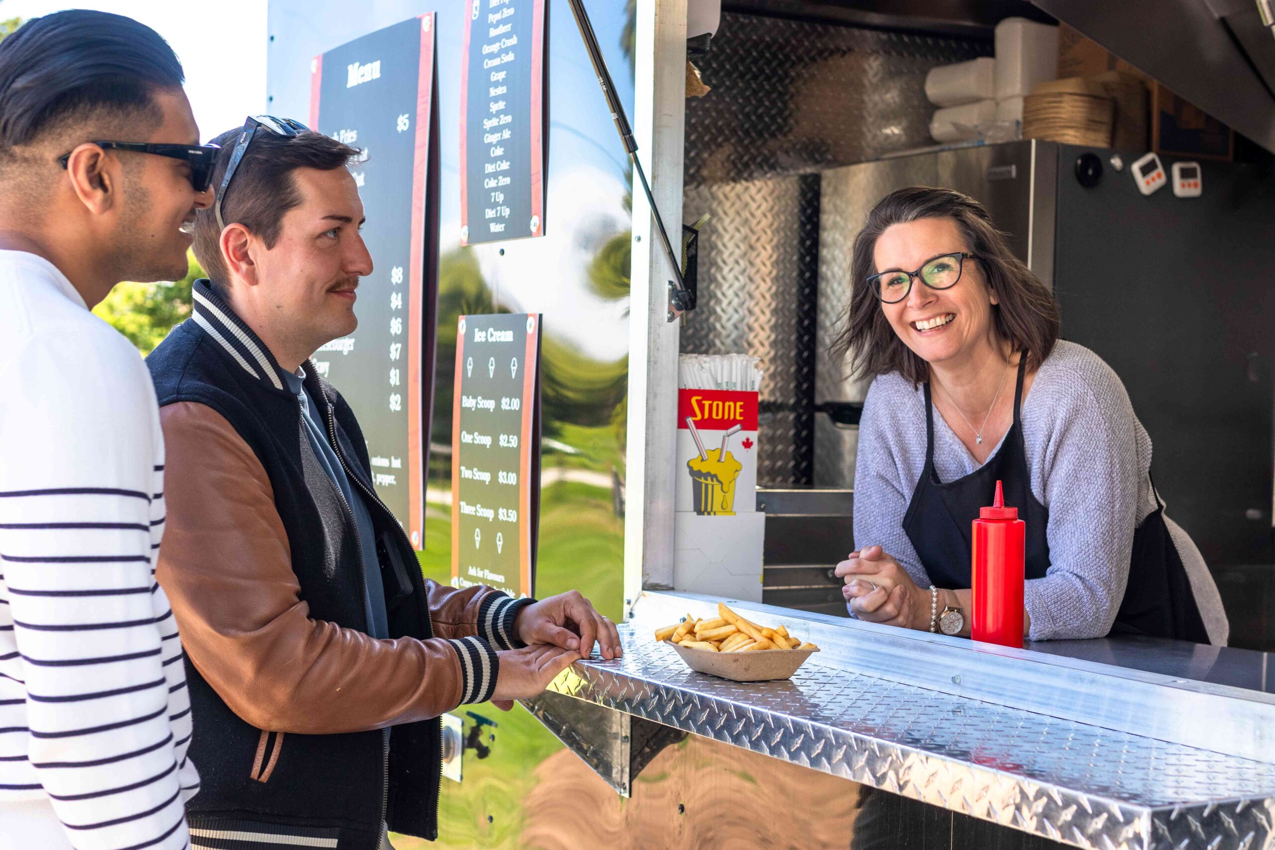 A woman smiles from a food truck service window while serving two customers.