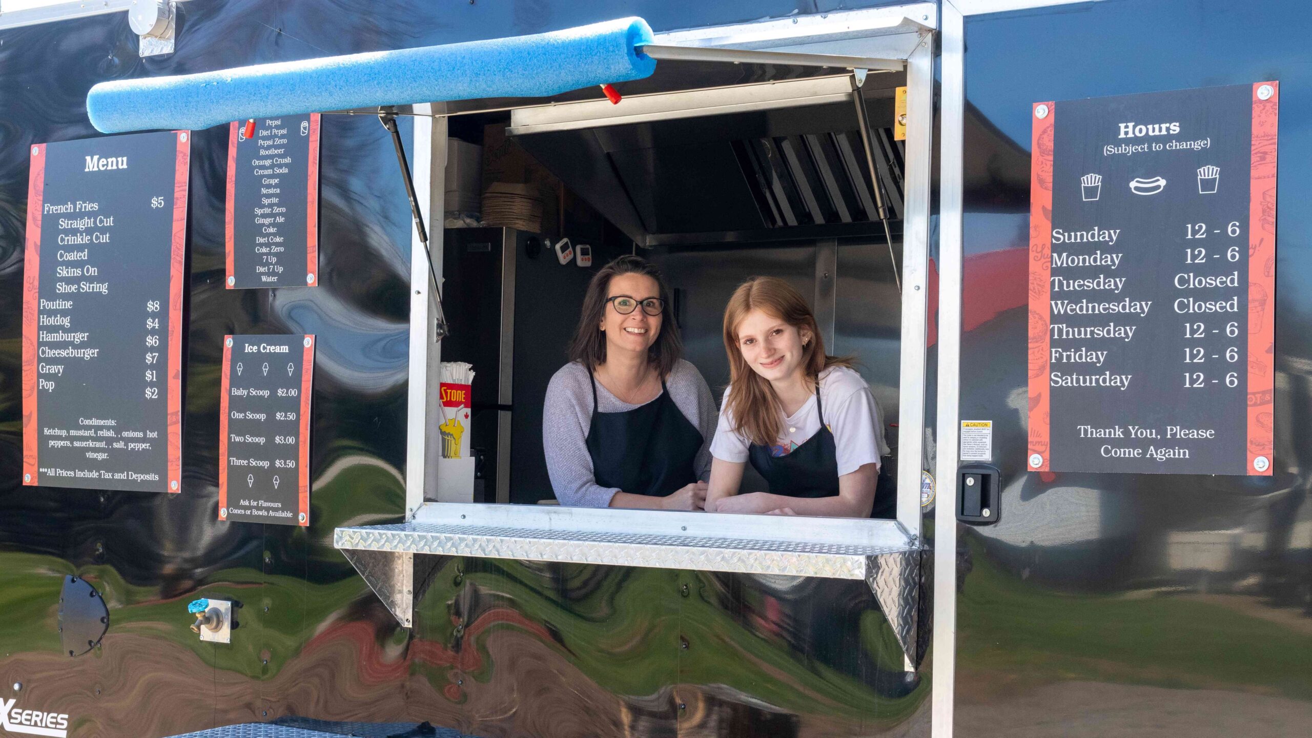Two women smile from the service window of a black food truck.